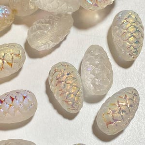 May include: A collection of iridescent glass pine cone shaped beads. The beads are white with a rainbow sheen.