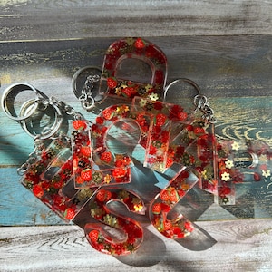 May include: Clear resin keychains spelling out the word "SHOP". Each letter is filled with red strawberries and gold glitter. The keychains are attached to silver keyrings, resting on a wooden surface.