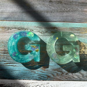 Pu&ograve; includere: Due ornamenti a forma di lettera "G" in resina. Uno &egrave; di un vivace verde acqua con accenti viola e oro incorporati. L'altro &egrave; verde traslucido con abbellimenti simili. Entrambi sono esposti su una superficie di legno invecchiato, colorata di blu.