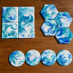 May include: A collection of ceramic coasters and tiles with a fluid art design. The coasters are round, and the tiles are square and hexagonal. The color palette includes shades of blue, teal, and white, creating a marbled effect.