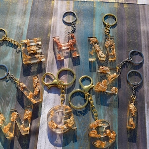 May include: A set of gold and clear resin letter keychains with gold leaf accents. The letters are W, N, G, S, H, and M. The keychains are on a wooden background.