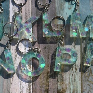 Pu&ograve; includere: Portachiavi con le lettere J, K, L, M, N, O, P e R. Ogni lettera &egrave; realizzata in resina trasparente con glitter iridescenti e piccole gemme. I portachiavi hanno anelli e catene in metallo argentato. Lo sfondo &egrave; una superficie di legno invecchiato.