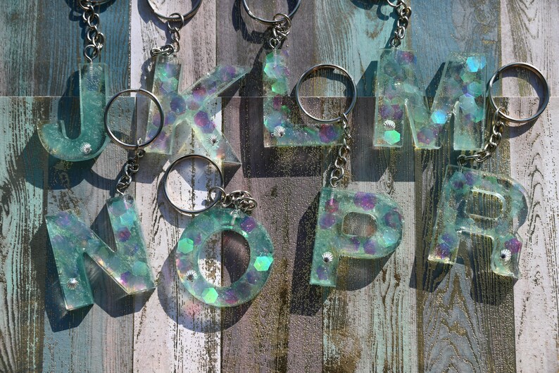 Pu&ograve; includere: Portachiavi con le lettere J, K, L, M, N, O, P e R. Ogni lettera &egrave; realizzata in resina trasparente con glitter iridescenti e piccole gemme. I portachiavi hanno anelli e catene in metallo argentato. Lo sfondo &egrave; una superficie di legno invecchiato.