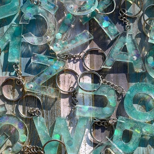 Pu&ograve; includere: Una collezione di portachiavi con lettere in resina trasparente in vari font. Le lettere sono riempite di glitter turchese e iridescenti, attaccate a portachiavi argentati. Lo sfondo &egrave; una superficie di legno invecchiato.