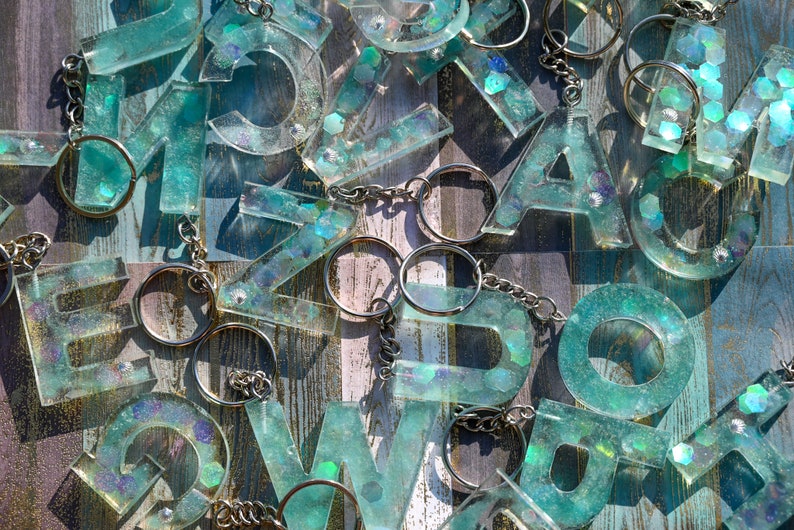 Pu&ograve; includere: Una collezione di portachiavi con lettere in resina trasparente in vari font. Le lettere sono riempite di glitter turchese e iridescenti, attaccate a portachiavi argentati. Lo sfondo &egrave; una superficie di legno invecchiato.