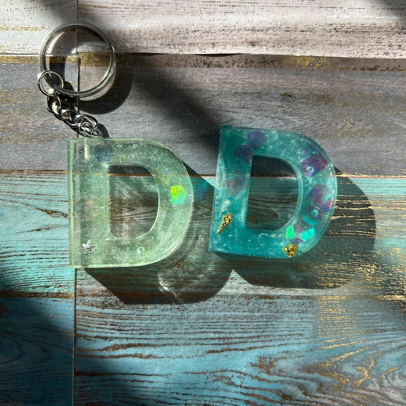 Pu&ograve; includere: Due portachiavi con la lettera "D" in resina. Uno &egrave; di colore verde chiaro, l'altro turchese. Entrambe le lettere hanno elementi decorativi all'interno, tra cui glitter e piccole forme. I portachiavi sono attaccati a un portachiavi argentato.