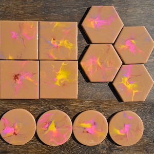 May include: A selection of decorative coasters in square, hexagonal, and round shapes. Each coaster has a tan base with colourful abstract designs in pink, yellow, and purple. The coasters are displayed on a wooden surface.
