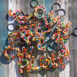 Puede incluir: Una colección de llaveros de letras coloridas hechos de resina transparente llena de chispas multicolores. Los llaveros están dispuestos en una pila y todos están unidos a llaveros plateados.