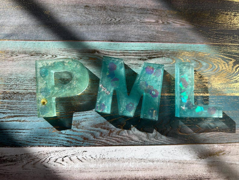 Pu&ograve; includere: Lettere in resina turchese che compongono "PML" con elementi decorativi incorporati. Le lettere sono su una superficie di legno con toni blu, grigi e oro. La lettera "P" ha un piccolo accento a conchiglia.
