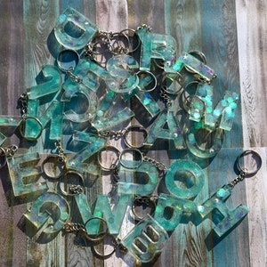 Pu&ograve; includere: Una collezione di portachiavi con lettere in resina turchese con portachiavi argentati. Ogni lettera &egrave; riempita di glitter iridescenti, creando un effetto scintillante. I portachiavi sono sparsi su una superficie di legno con una combinazione di colori blu e grigio.