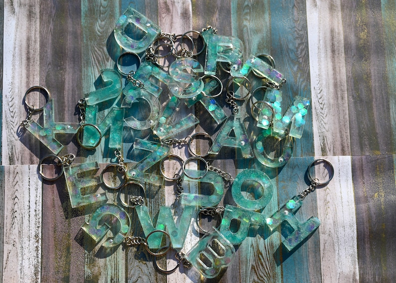 Pu&ograve; includere: Una collezione di portachiavi con lettere in resina turchese con portachiavi argentati. Ogni lettera &egrave; riempita di glitter iridescenti, creando un effetto scintillante. I portachiavi sono sparsi su una superficie di legno con una combinazione di colori blu e grigio.