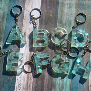 Pu&ograve; includere: Portachiavi con le lettere dalla A alla H, realizzati in resina trasparente con un effetto glitter turchese e iridescente. Ogni lettera &egrave; attaccata a un portachiavi e a una catena color argento. I portachiavi sono esposti su una superficie di legno.