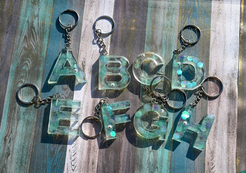 Pu&ograve; includere: Portachiavi con le lettere dalla A alla H, realizzati in resina trasparente con un effetto glitter turchese e iridescente. Ogni lettera &egrave; attaccata a un portachiavi e a una catena color argento. I portachiavi sono esposti su una superficie di legno.