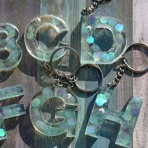 Pu&ograve; includere: Una collezione di portachiavi con lettere in resina trasparente, ciascuno con catena e anello argentati. Le lettere sono riempite di glitter iridescenti e piccoli dettagli a conchiglia, creando un effetto scintillante. Lettere: B, C, D, F, G, H.
