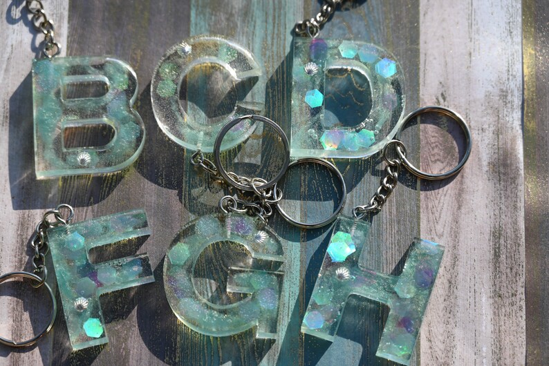 Pu&ograve; includere: Una collezione di portachiavi con lettere in resina trasparente, ciascuno con catena e anello argentati. Le lettere sono riempite di glitter iridescenti e piccoli dettagli a conchiglia, creando un effetto scintillante. Lettere: B, C, D, F, G, H.