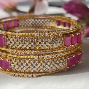 May include: A set of three gold bangles featuring pink and white stones. The bangles are adorned with a repeating pattern of small white stones and larger pink stones.