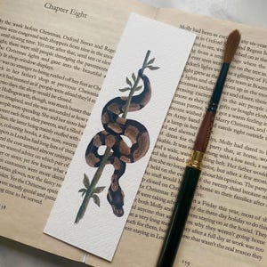 May include: Watercolor bookmark featuring a brown and tan snake coiled around a green stem. The bookmark is rectangular and rests on a book page with text. A paintbrush is next to the bookmark.