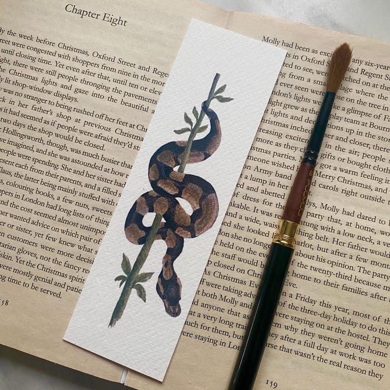 May include: A watercolour bookmark depicting a brown and tan snake wrapped around a green stem. The bookmark is rectangular and lies on a book page. A paintbrush is positioned nearby.