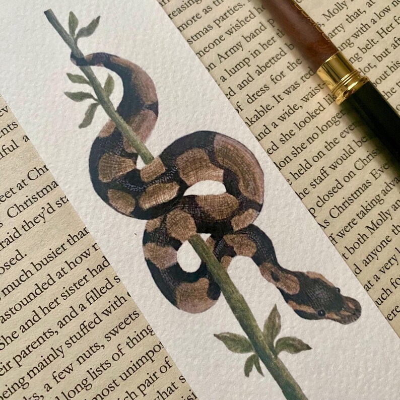 May include: A watercolour painting of a brown and black snake wrapped around a green branch. The snake's scales are detailed, set against a backdrop of vintage book pages. A pen is in the upper right corner.