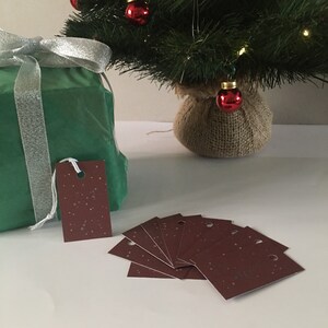 Peut inclure: Un ensemble de dix étiquettes cadeaux brunes avec un motif blanc moucheté et un trou pour la ficelle. Les étiquettes sont disposées en éventail sur une surface blanche.