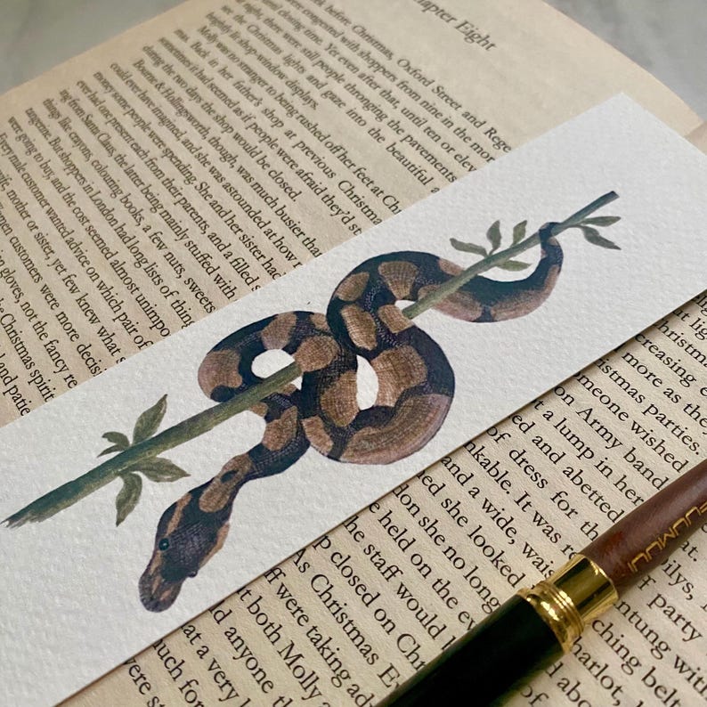 May include: A watercolour bookmark depicting a brown and black snake wrapped around a green branch. The snake's markings are detailed. The bookmark is set against a backdrop of book pages.