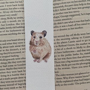 May include: Watercolor bookmark featuring a detailed illustration of a hamster. The hamster is depicted in shades of brown and tan, with dark ears and a focused expression. The bookmark is rectangular and set against a backdrop of printed text.