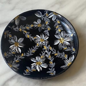 May include: A black ceramic dish with a white and gold floral design. The dish is decorated with white flowers and gold leaves on a black background.