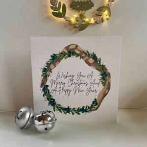 Peut inclure: Une carte de Noël carrée avec un motif de couronne avec un motif de serpent et du feuillage vert. La carte porte le texte "Wishing You A Merry Christmas And A Happy New Year". Deux cloches argentées sont au premier plan.