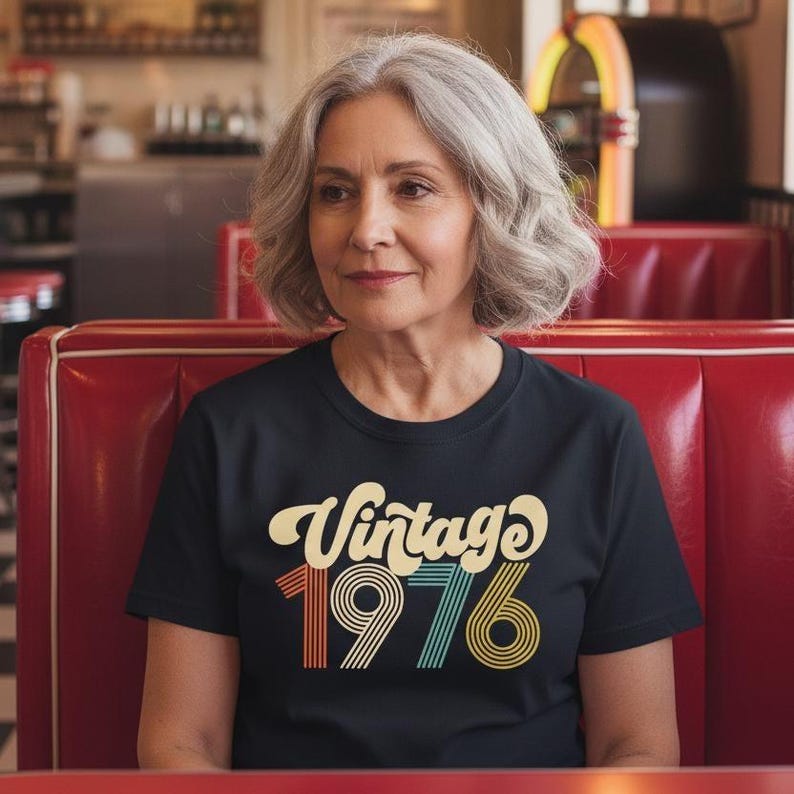 Peut inclure: Une femme porte un t-shirt noir avec l'inscription "Vintage 1976" dans une police r&eacute;tro. Le texte est de couleur cr&egrave;me, orange et turquoise. La femme est assise dans une cabine rouge dans un cadre de restaurant.