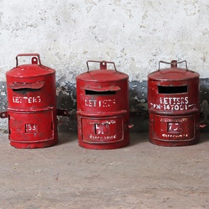 May include: Three red vintage mailboxes with white lettering. The mailboxes have two compartments, one for letters and one for clearance. The top compartment has the word "LETTERS" written on it. The bottom compartment has a number on it. The mailboxes are sitting on a concrete floor.