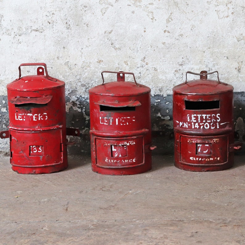 May include: Three red vintage mailboxes with white lettering. The mailboxes have two compartments, one for letters and one for clearance. The top compartment has the word "LETTERS" written on it. The bottom compartment has a number on it. The mailboxes are sitting on a concrete floor.