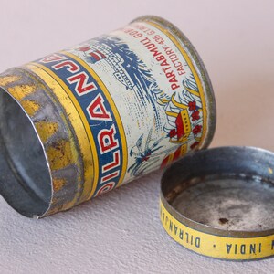 May include: Vintage metal tin with a cylindrical shape, featuring a yellow and blue label with text and illustrations. The lid is separate, showing the words "MADE IN INDIA". The tin is slightly dented, showing signs of age.