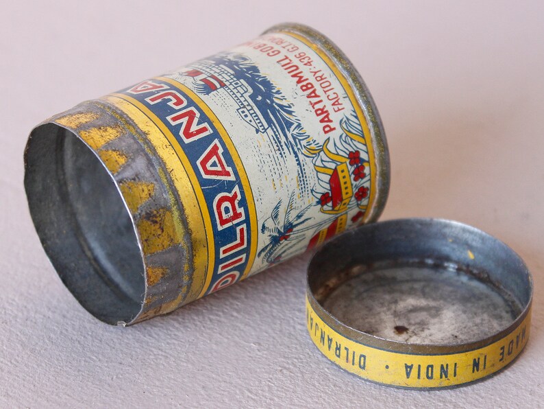 May include: Vintage metal tin with a cylindrical shape, featuring a yellow and blue label with text and illustrations. The lid is separate, showing the words "MADE IN INDIA". The tin is slightly dented, showing signs of age.