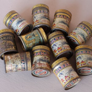 May include: A collection of vintage metal tins, featuring the text "DILRANJA" and other inscriptions. The tins have a distressed, aged appearance with illustrations of buildings and palm trees. The lids are gold-colored.