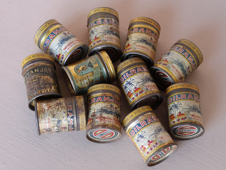 May include: A collection of vintage metal tins, featuring the text "DILRANJA" and other inscriptions. The tins have a distressed, aged appearance with illustrations of buildings and palm trees. The lids are gold-colored.
