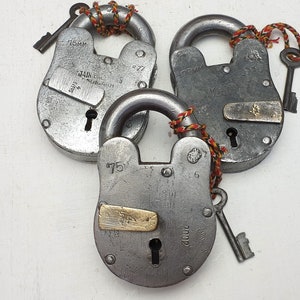 Könnte beinhalten: Drei antike silberne Vorhängeschlösser mit Schlüsseln. Jedes Vorhängeschloss hat eine andere Größe und einen anderen Markennamen. Die Vorhängeschlösser sind mit roter und gelber Schnur zusammengebunden.