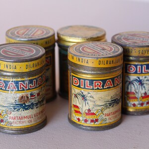 May include: A collection of vintage metal tins with yellow, blue, and red labels. The labels feature the text "DILRANJAN" and "MADE IN INDIA". The tins have a cylindrical shape and show signs of age.