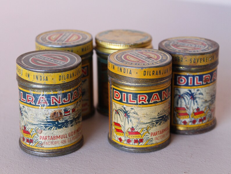 May include: A collection of vintage metal tins with yellow, blue, and red labels. The labels feature the text "DILRANJAN" and "MADE IN INDIA". The tins have a cylindrical shape and show signs of age.