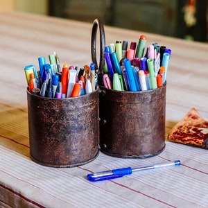 Può includere: Due contenitori in metallo rustico pieni di una varietà di penne e matite colorate. Una penna blu giace su una superficie di tavolo a righe.