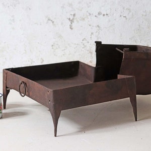 Può includere: Un braciere in metallo arrugginito con quattro gambe e una base quadrata. Il braciere è in metallo e ha un aspetto rustico. È appoggiato su una superficie bianca.