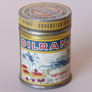 May include: Vintage metal tin with a yellow and blue label. The label features the word "DILRANJAN" in red letters, along with illustrations of palm trees, buildings, and a ship. The tin has a gold-colored lid with text.