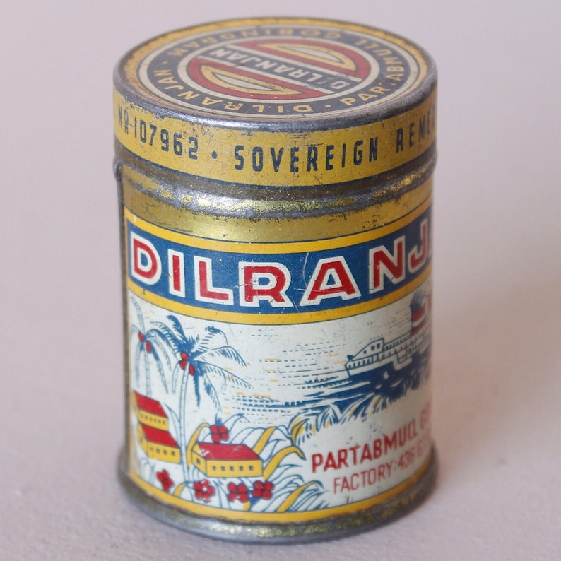 May include: Vintage metal tin with a yellow and blue label. The label features the word "DILRANJAN" in red letters, along with illustrations of palm trees, buildings, and a ship. The tin has a gold-colored lid with text.