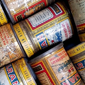 May include: A collection of vintage metal tins with faded labels. The tins feature text in multiple languages, with red, blue, and yellow color schemes. Some labels include the text "SOVEREIGN REMEDY" and "ALGOBINDRAM".