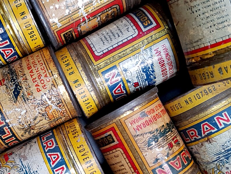 May include: A collection of vintage metal tins with faded labels. The tins feature text in multiple languages, with red, blue, and yellow color schemes. Some labels include the text "SOVEREIGN REMEDY" and "ALGOBINDRAM".