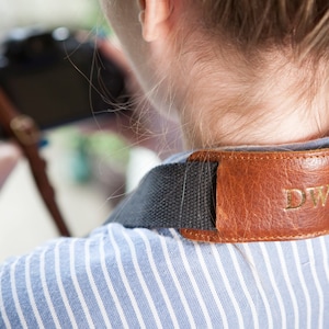 Può includere: Una tracolla per fotocamera in pelle marrone con le iniziali "DW" impresse in oro. La tracolla è attaccata a una fotocamera nera e la persona indossa una camicia a righe blu e bianche.