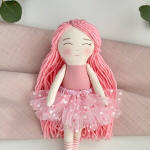 Puede incluir: Mu&ntilde;eca de tela hecha a mano con pelo de hilo rosa, un vestido rosa claro y un tut&uacute; rosa con lunares blancos y rosas. La mu&ntilde;eca tiene una cara sonriente y calcetines a rayas. Es un juguete infantil encantador.