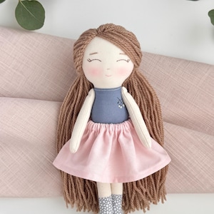 Peut inclure: Poup&eacute;e en tissu faite &agrave; la main avec de longs cheveux bruns en fil, un haut bleu et une jupe rose. La poup&eacute;e a un visage doux et souriant avec des joues roses et des chaussettes grises &agrave; motifs. Un article charmant et artisanal.