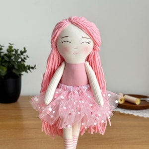 Puede incluir: Mu&ntilde;eca de tela hecha a mano con pelo de hilo rosa y una cara sonriente. La mu&ntilde;eca lleva un maillot rosa, un tut&uacute; con lunares blancos y calcetines a rayas. Un juguete encantador y caprichoso.