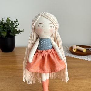 May include: A handmade fabric doll with long, light blonde yarn hair and a serene expression. The doll wears a blue polka-dot top, an orange skirt, and matching orange boots. The doll is a charming example of handcrafted textile art.