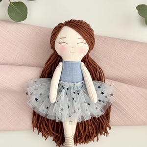 May include: A handmade doll with brown yarn hair, a blue denim bodice, and a white tulle skirt adorned with navy blue stars. The doll has a sweet, smiling face and is wearing striped socks. A charming, handcrafted toy.